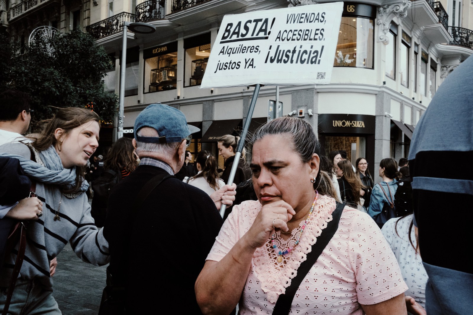 Taller de Inmersión en Fotografía de Calle en Madrid con Rober Tomás. Una mujer que pasa delante de un señor mayor que lleva una pancarta de protesta a favor de la Vivienda en España y una mujer que pasa al lado con actitud pensativa en Gran Vía