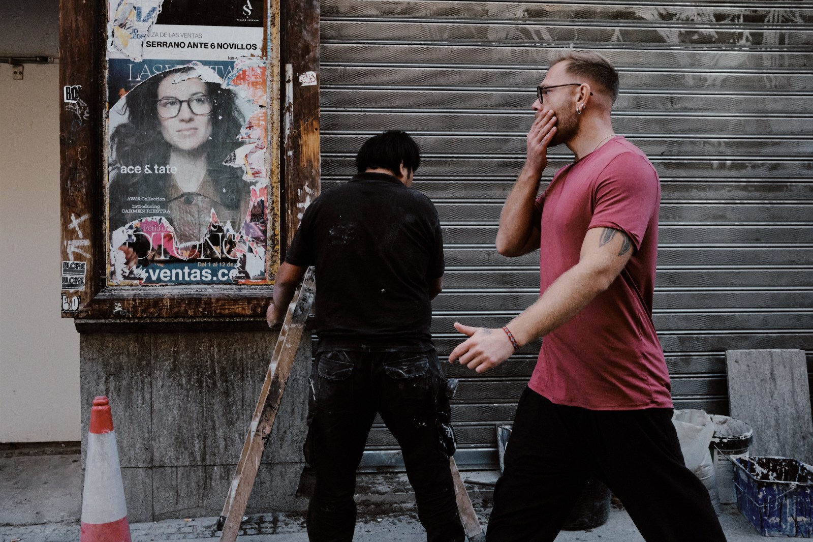Taller de Inmersión en Fotografía de Calle en Madrid con Rober Tomás. Personas pasando por la calle un cartel con una chica con gafas y un chico con camiseta roja y con gafas mirándola