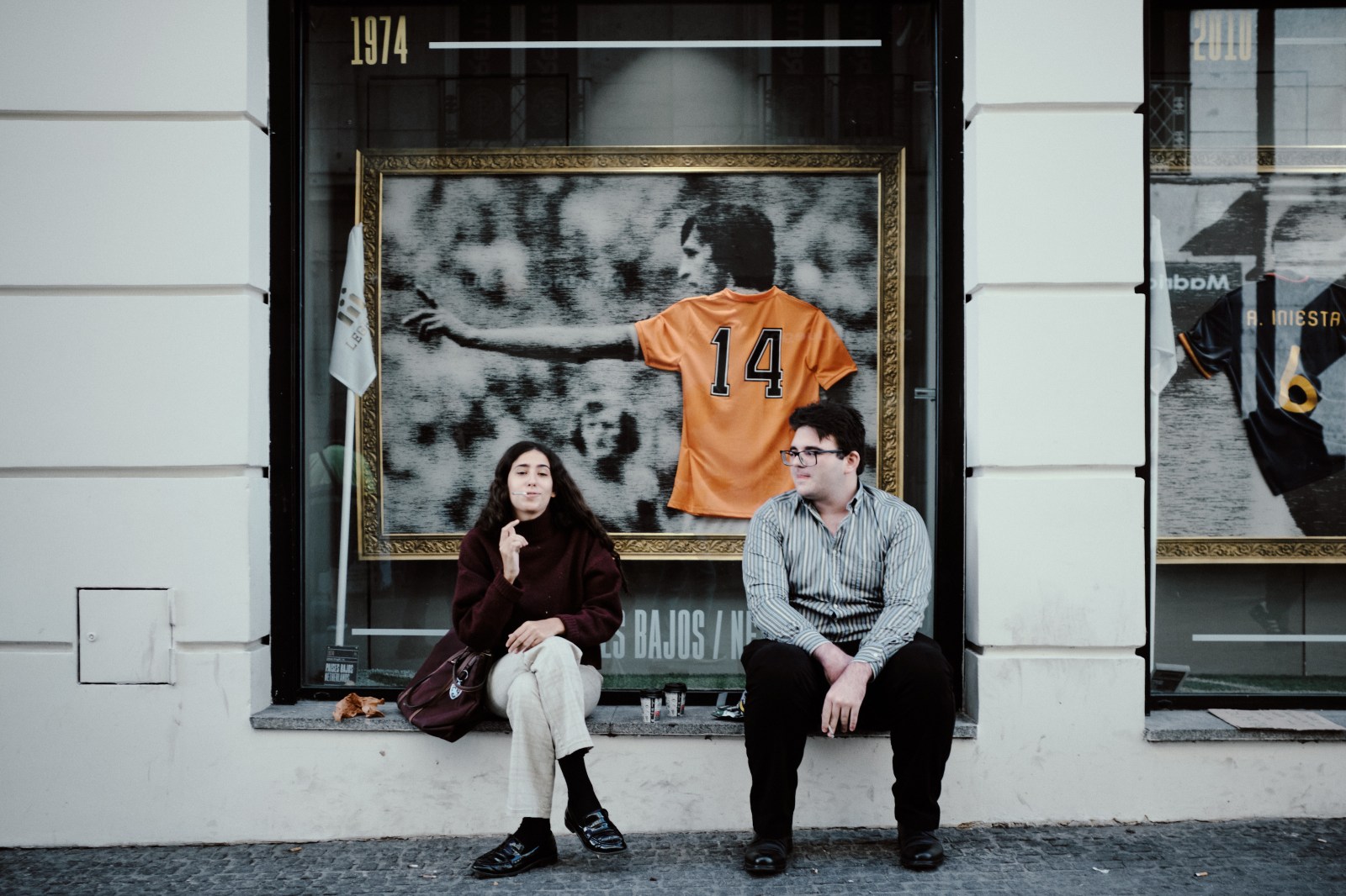 Taller de Inmersión en Fotografía de Calle en Madrid con Rober Tomás. Un chico y una chica sentados debajo de una fotografia de Johan Cruyff 