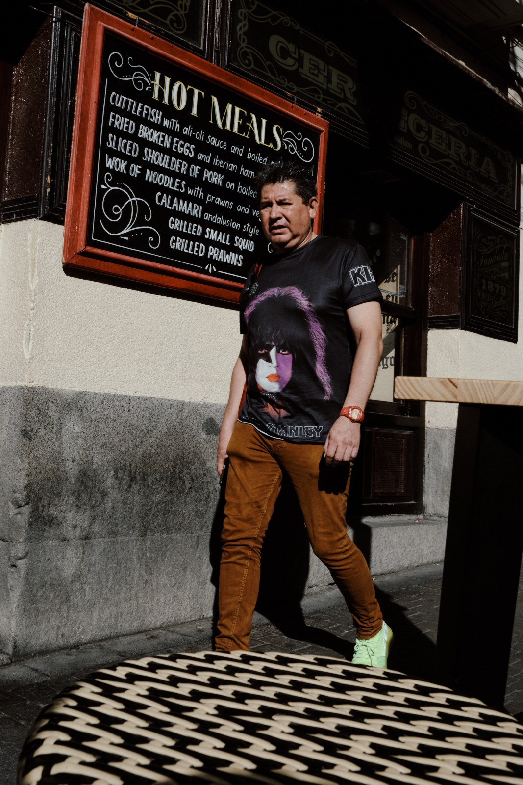 Taller de Inmersión en Fotografía de Calle en Madrid con Rober Tomás. Unhombre que pasa delante de un bar antiguo de Madrid con una camiseta del grupo de rock Kiss
